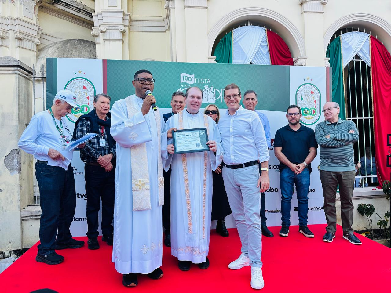 Entrega da documentação de compensação na 100ª Festa de Nossa Senhora Achiropita, em frente à Igreja Nossa Senhora Achiropita no Bixiga, São Paulo, com identificação visual da 100ª edição ao fundo